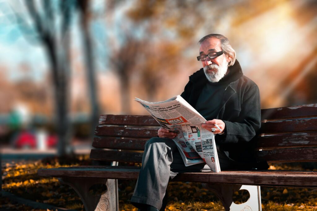 An older man wearing glasses sits on an outdoor bench and reads a newspaper. It is sunny outside and there are trees behind him.