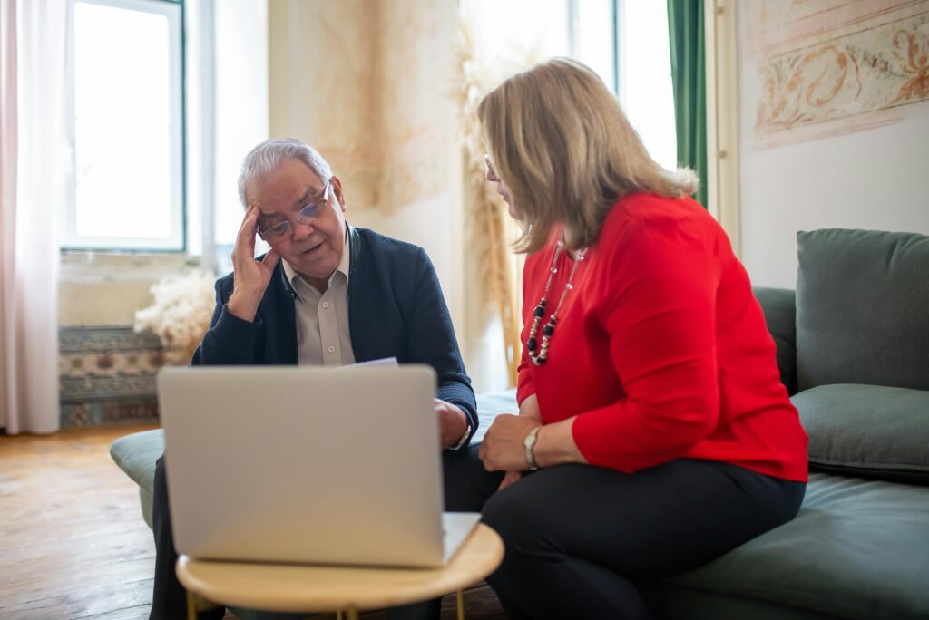 A man and a woman sit on a couch with a laptop open on a small table in front of them. Both look concerned.