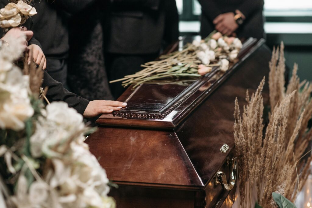 A closed wooden casket with white roses laid on top. A mourner lays their hand on the lid. In the foreground are white roses.