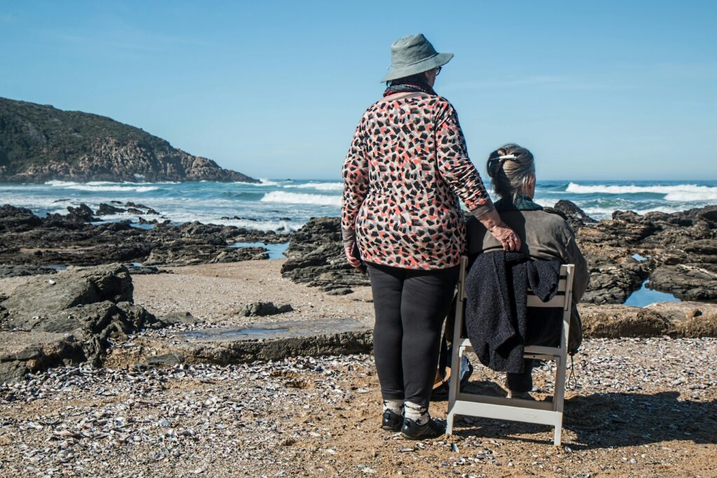 Woman Standing Beside Elderly Woman on White Wooden Chair Facing Body of Water