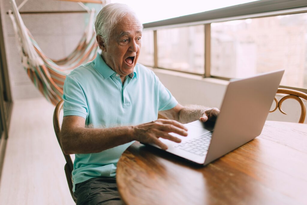 An older man wearing a blue shirt looks shocked while staring at his laptop screen.