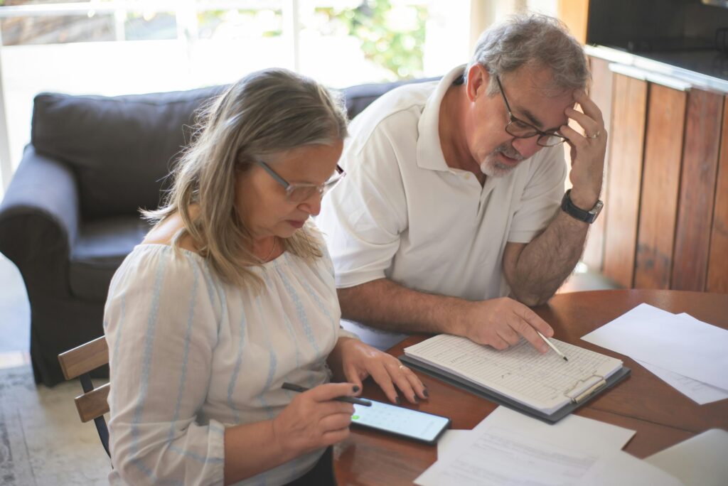 Senior couple reviewing documents together at a home table while using a smartphone.