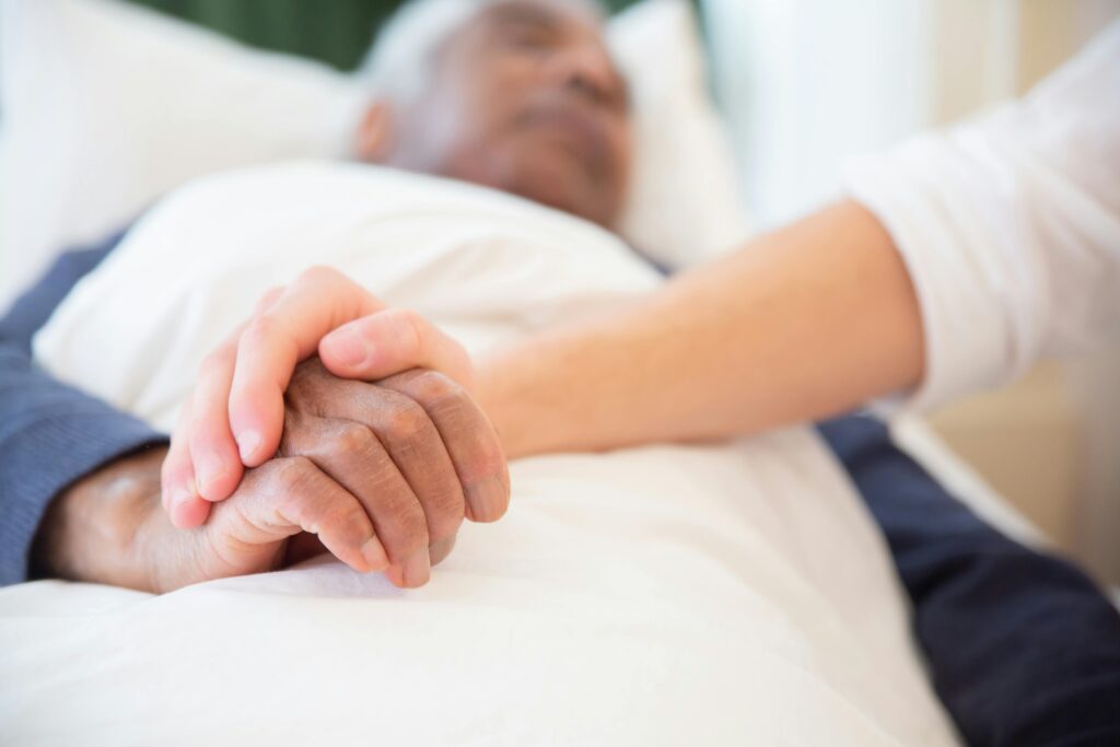 Up-close shot of a young person holding an elderly man's hand as he lays in a hospital bed.
