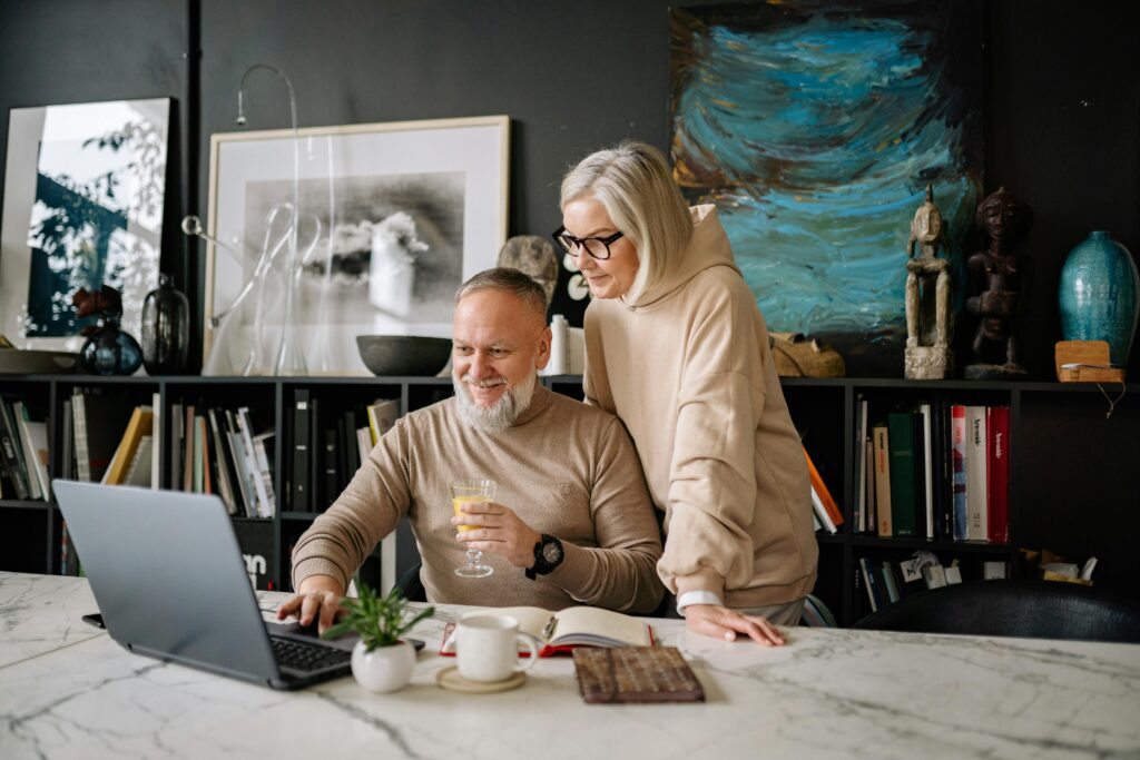 An older couple at a marble table, looking at a laptop screen. The woman stands, the man sits.