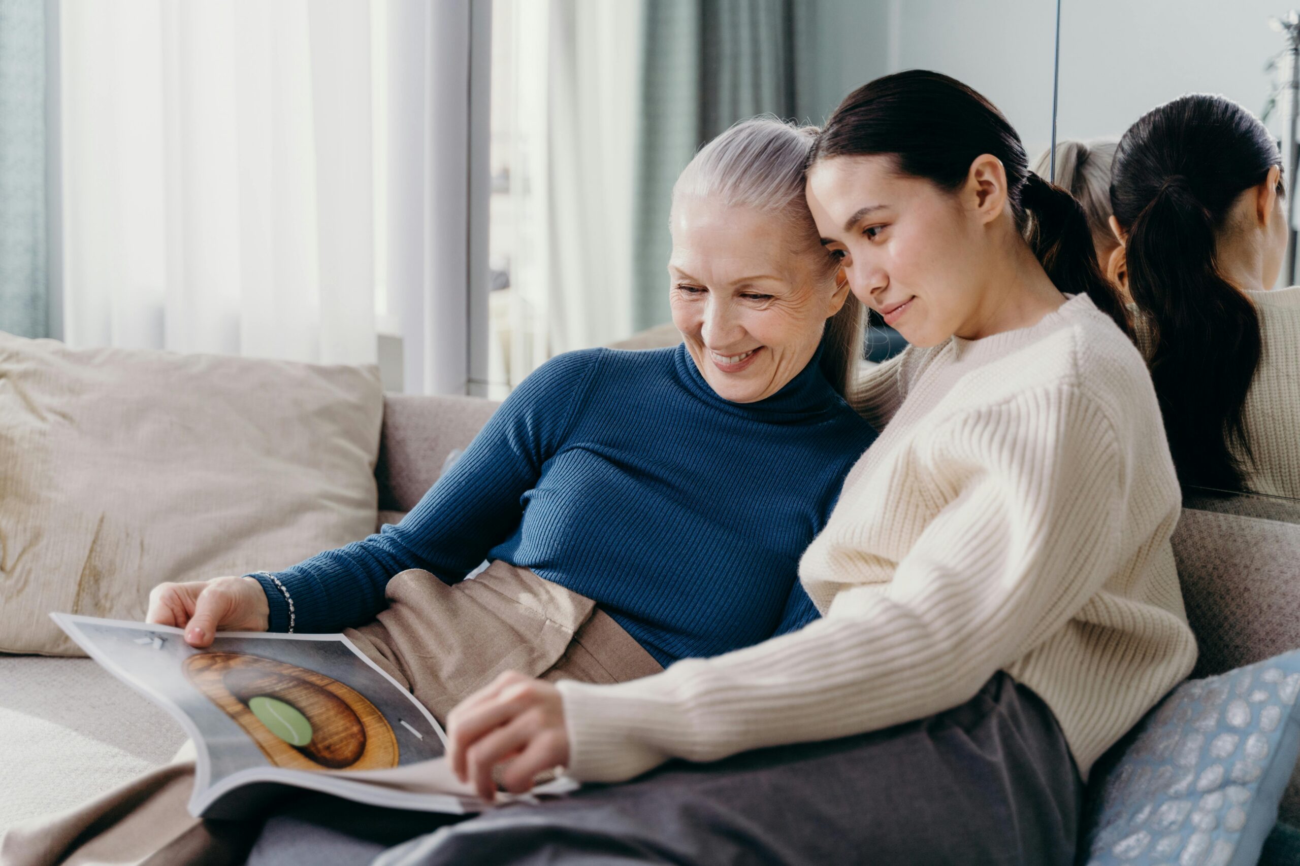 A mother and daughter are sitting on a couch, looking at a magazine.