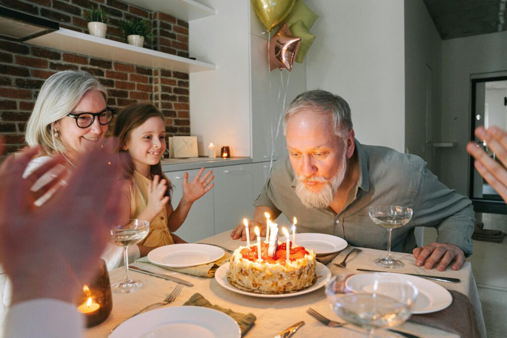 An older man blows out the candles on a birthday cake while family members surround him, clapping. Several pink and yellow star-shaped balloons hover in the background.
