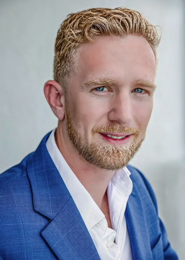 Head-and-shoulders portrait of a smiling man with blond hair and a trimmed beard in a blue blazer and white shirt, looking at the camera.