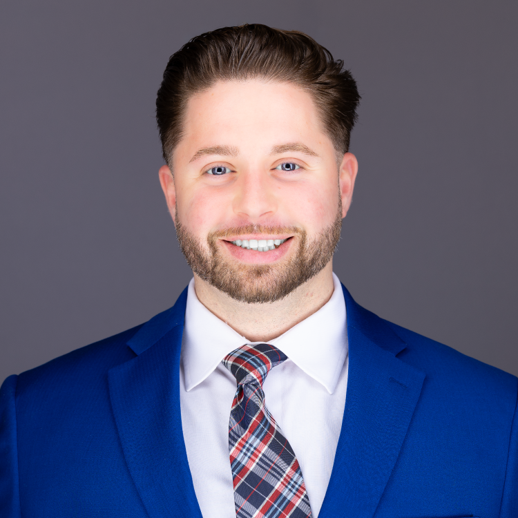 Professional headshot of a smiling man in a blue suit and plaid tie against a gray backdrop.