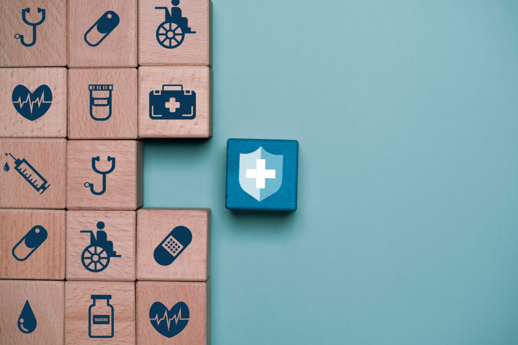 Healthcare insurance and medical protection concept with wooden blocks showing hospital, vaccine, syringe and wellness icons beside shield symbol.